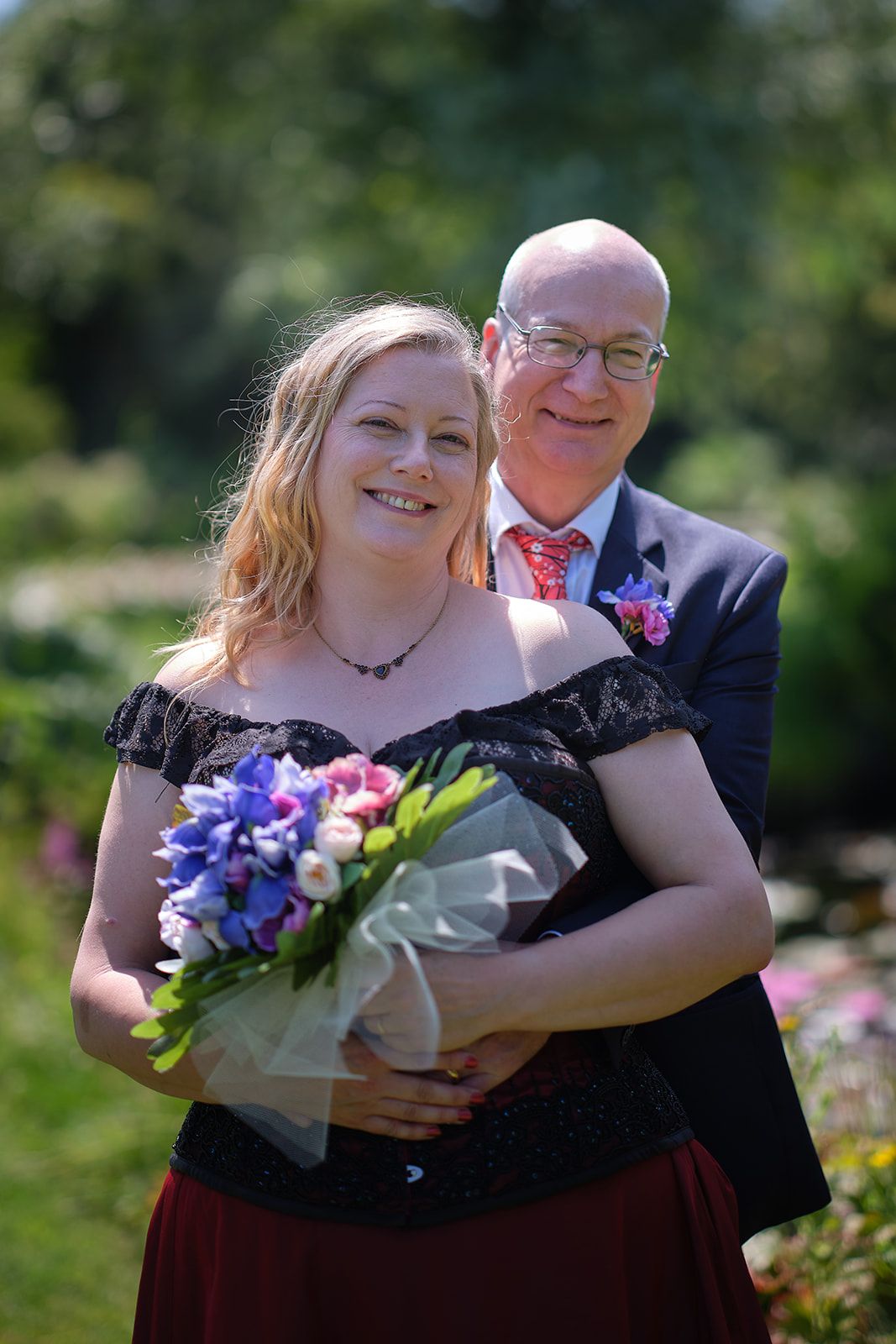 A couple poses together at an outdoor event, with the woman wearing a black dress and holding a colorful floral bouquet.