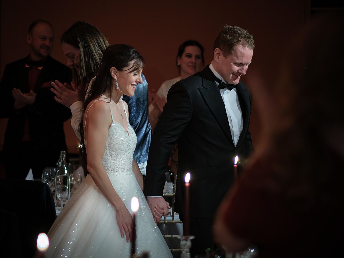 Bride in white gown and groom in black tuxedo smile at candlelit wedding reception dinner table.