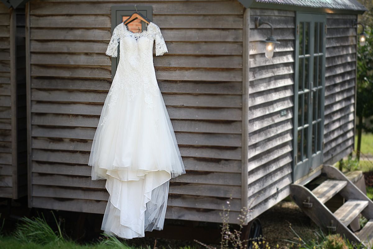 White lace wedding dress hangs elegantly against rustic wooden cabin siding with a window visible in the background.