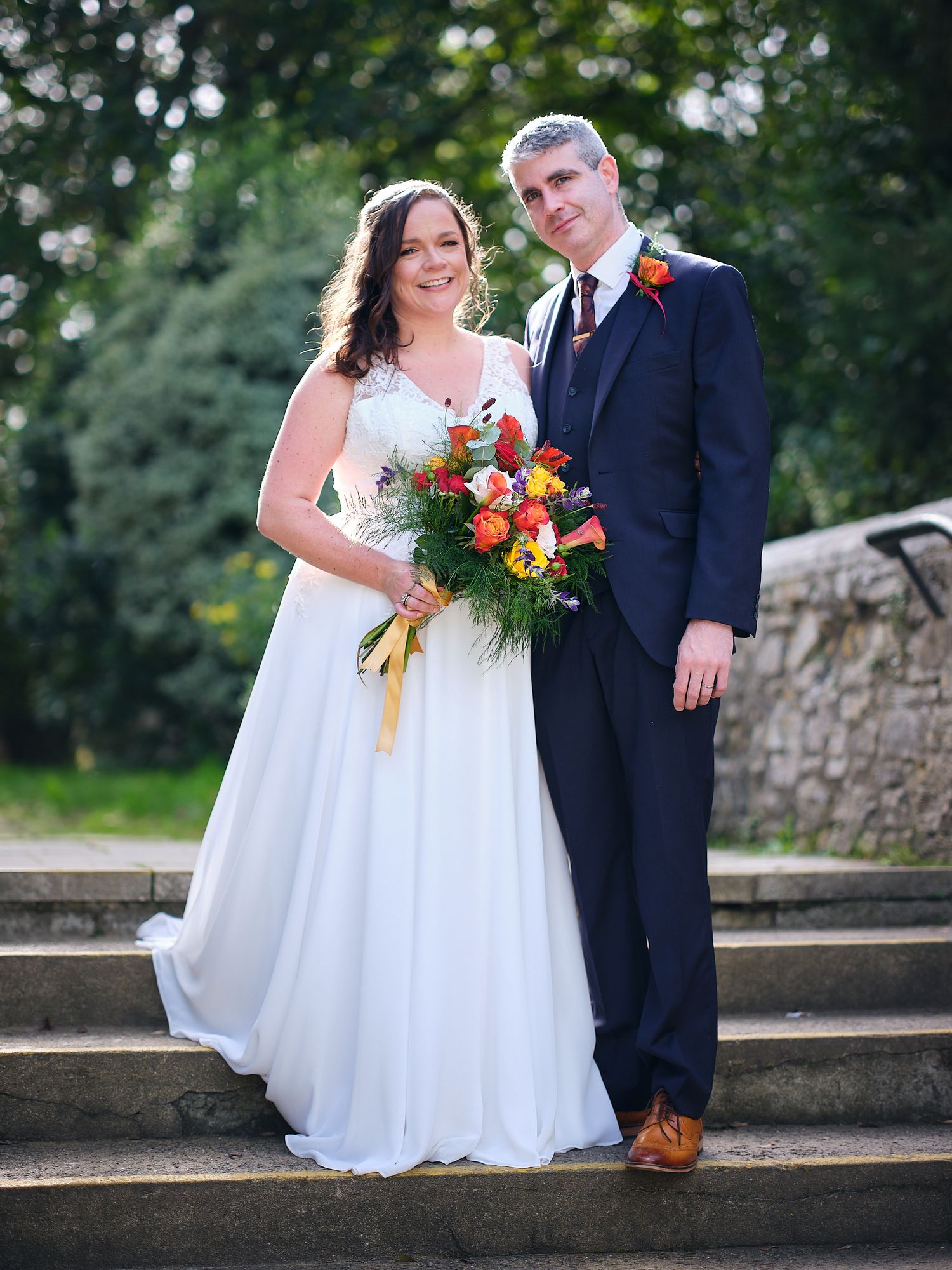 A couple in wedding attire poses on stone steps surrounded by greenery as the bride holds a vibrant autumn bouquet.