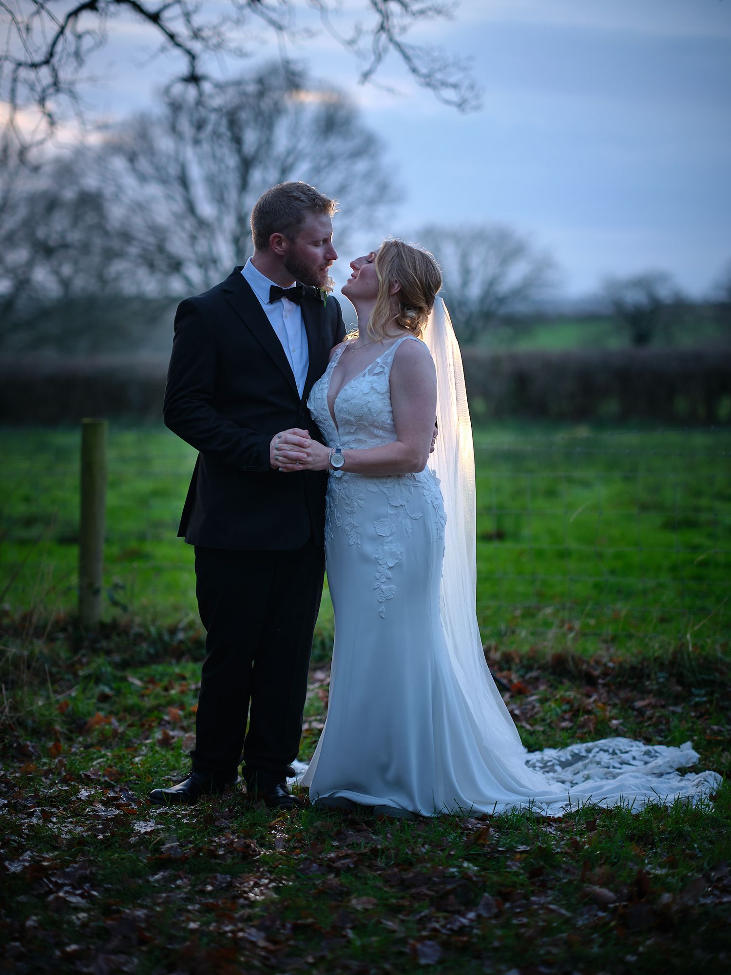 A romantic couple in formal wedding attire embraces in a serene outdoor setting with green fields at dusk.