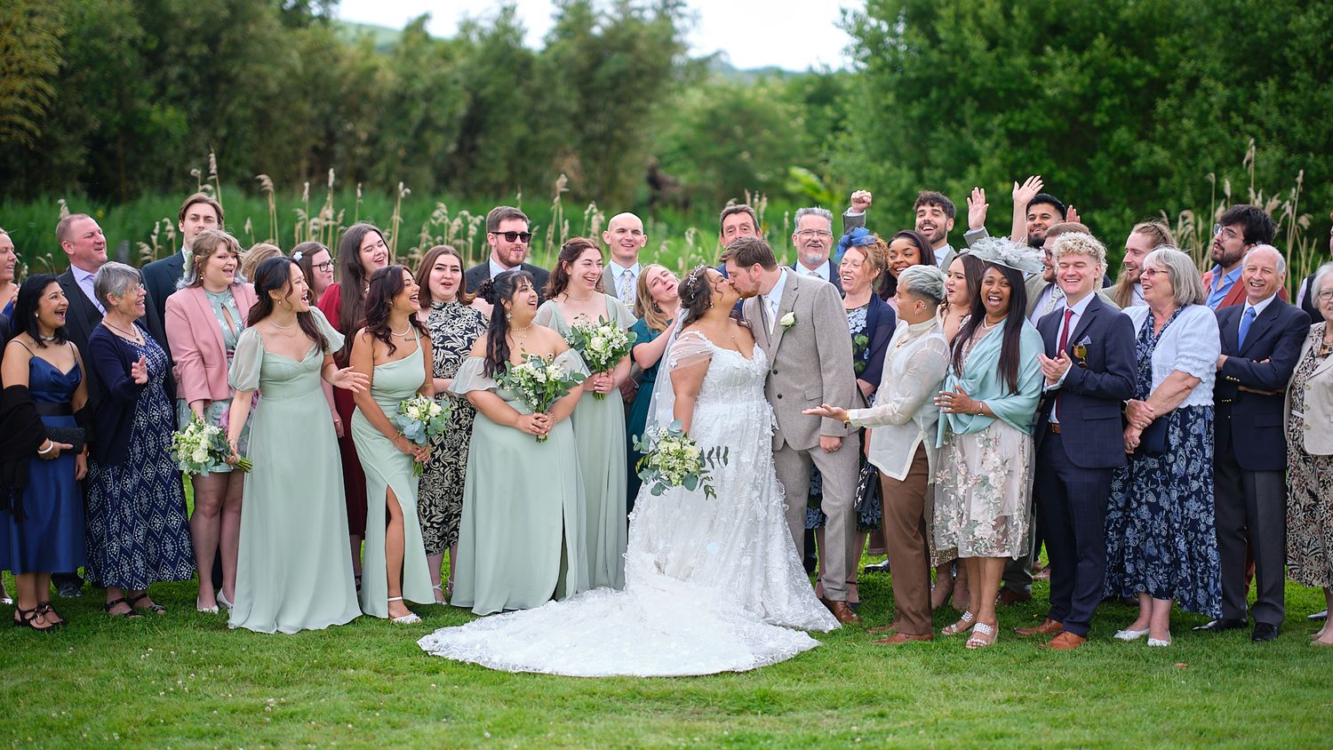 Wedding guests gather outdoors in elegant attire amidst green trees and landscaping during a sunny summer celebration.