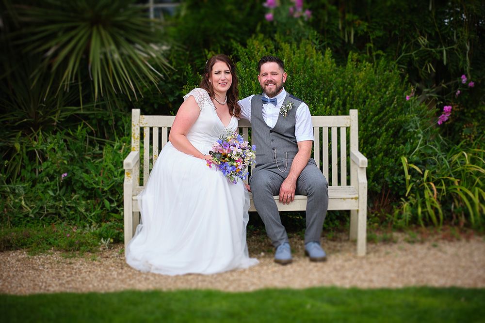 A couple sits together on a white garden bench surrounded by lush green plants and flowers.
