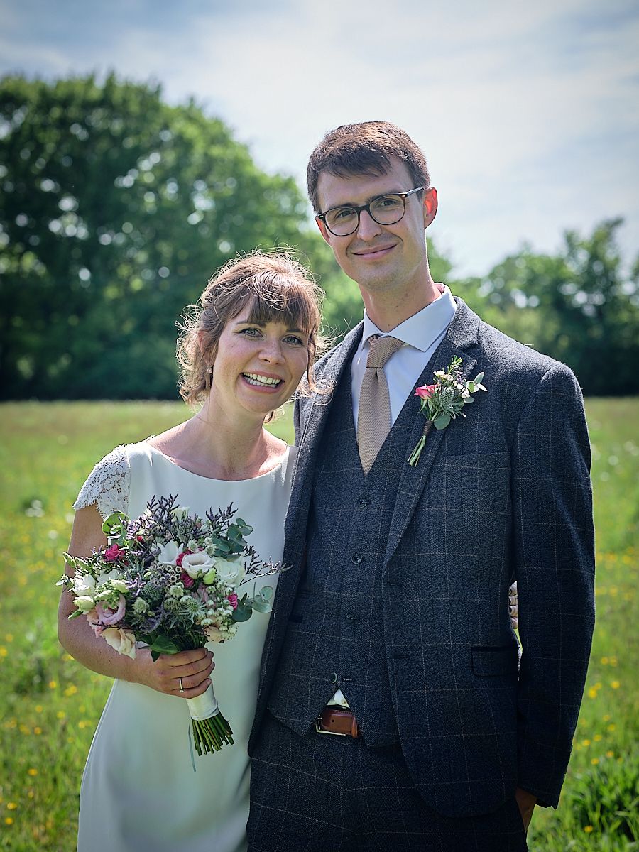 A couple in wedding attire pose together in a sunny meadow with greenery, holding a bouquet of pink and white flowers.