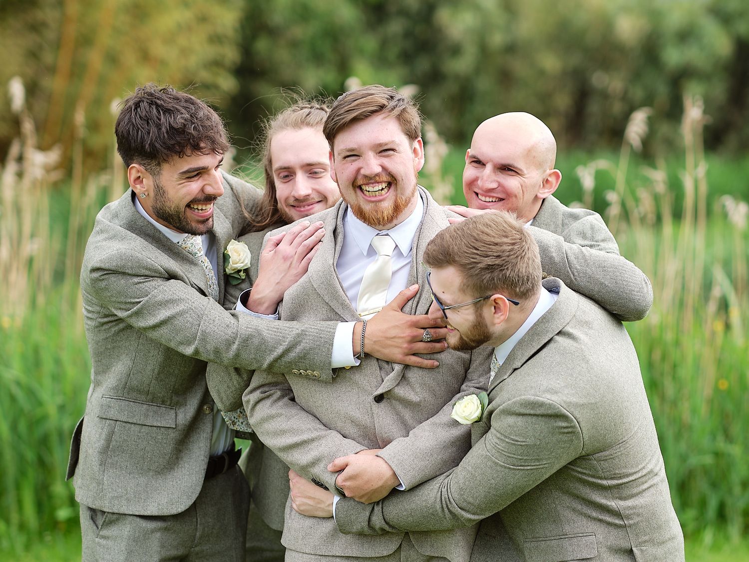 Four groomsmen wearing matching gray suits laugh and embrace each other in a playful moment outdoors in nature.
