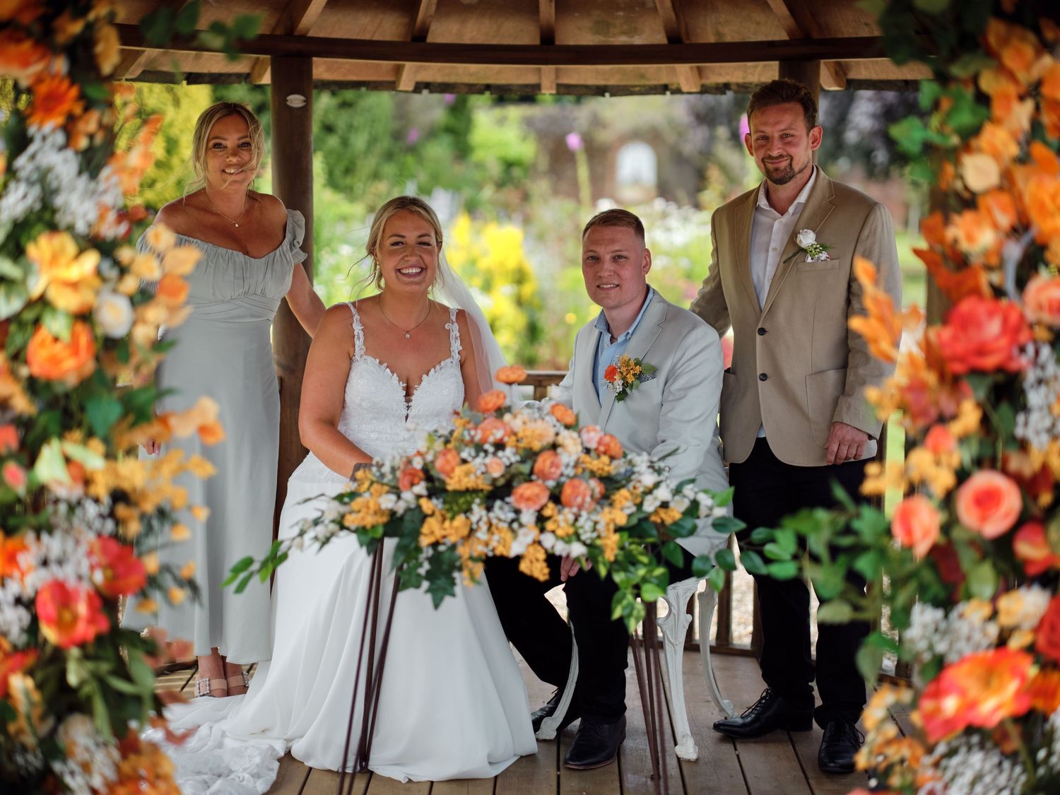 Wedding party poses under a beautiful pergola decorated with vibrant orange and peach floral arrangements.
