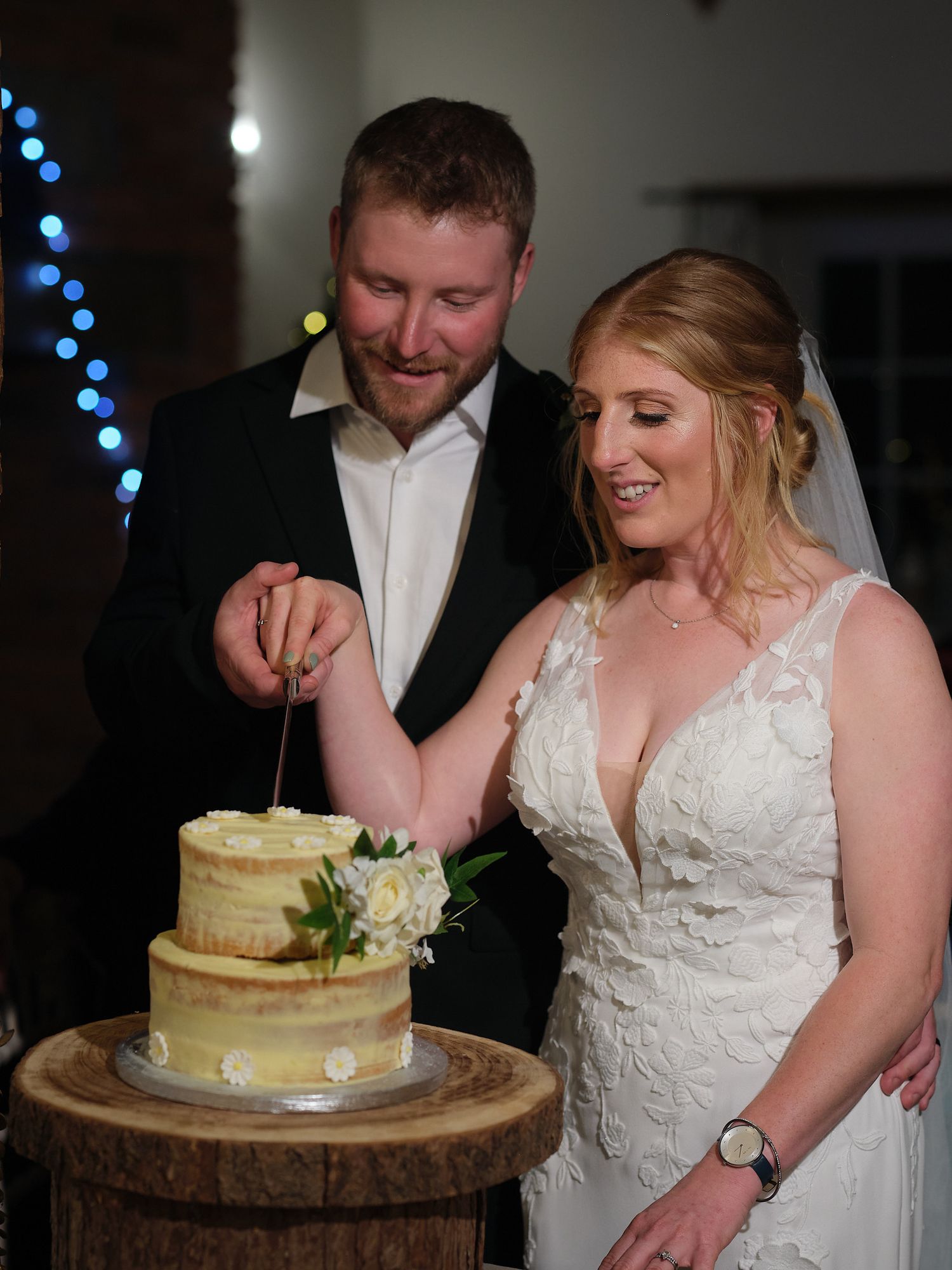 A bride and groom cut into their two-tiered semi-naked wedding cake decorated with greenery on a rustic wooden stand.