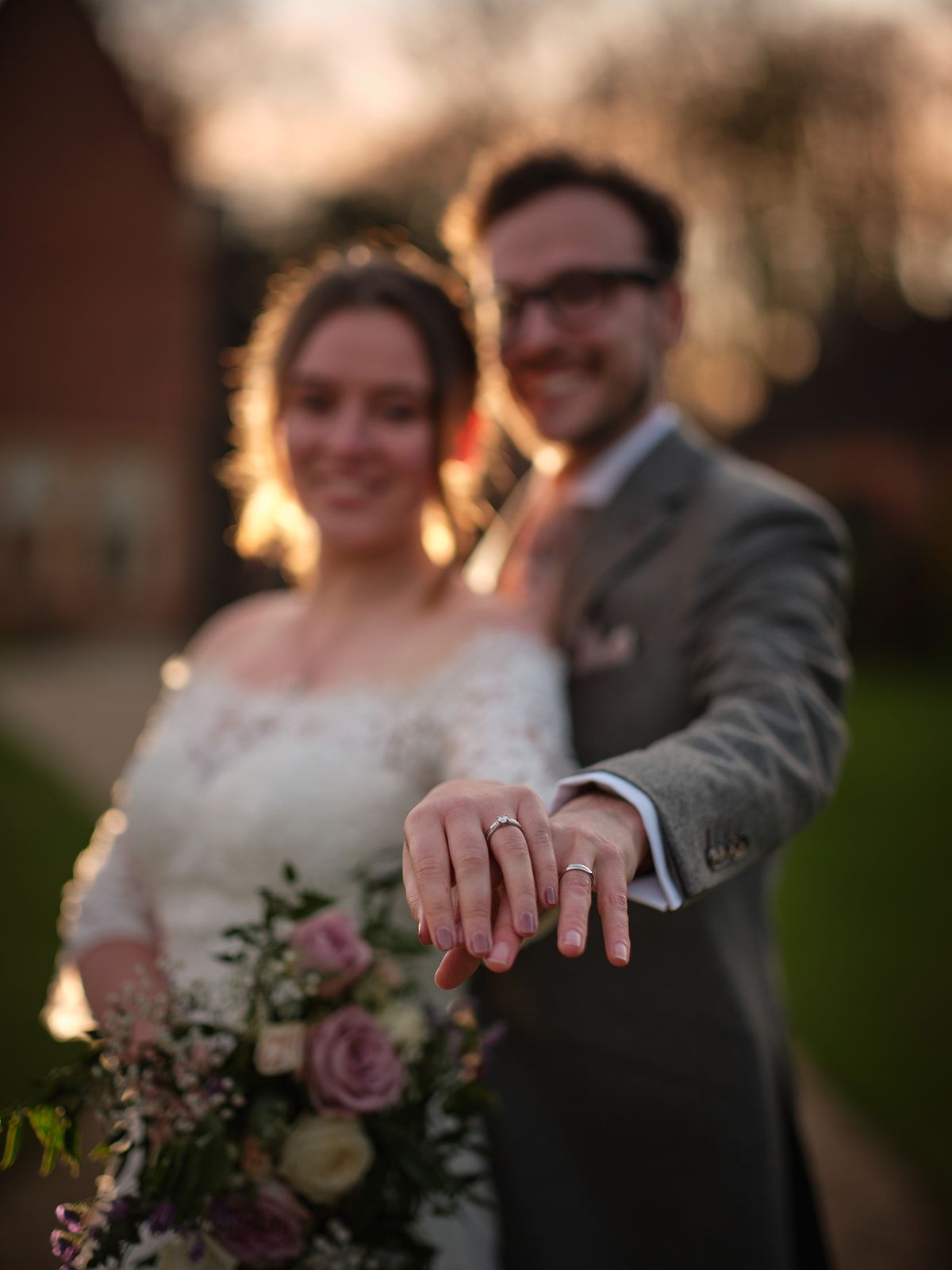 Newlywed couple showing off wedding rings in golden sunset light with blurred background and bridal bouquet.