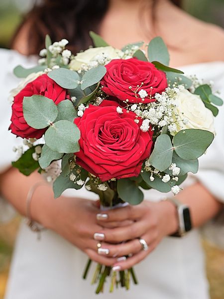 A bridal bouquet featuring vibrant red roses, white baby's breath, and soft eucalyptus leaves held by manicured hands.
