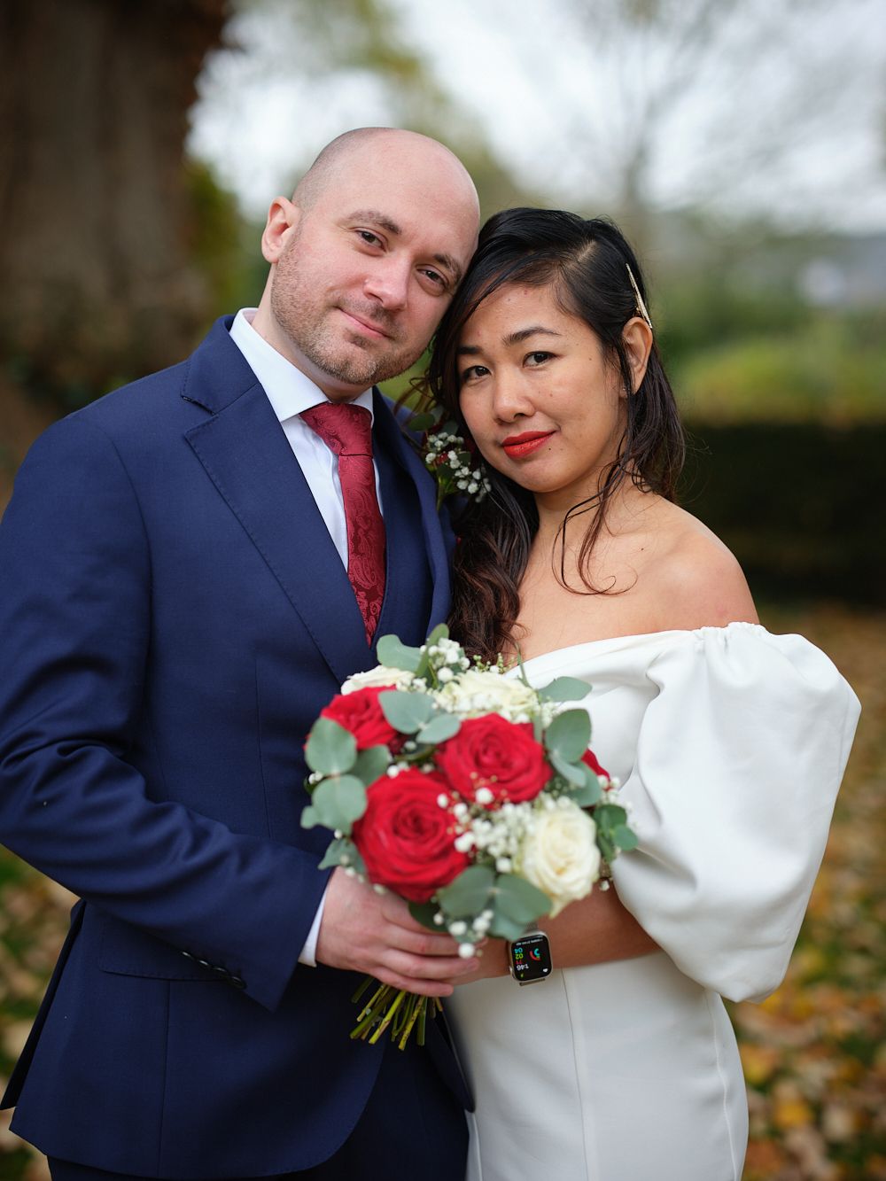 A bride and groom pose together outdoors, with the bride holding a bouquet of red and white roses with greenery.