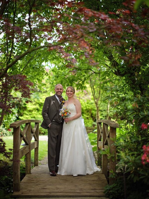 A couple stands on a wooden footbridge surrounded by lush Japanese maple trees and greenery in a garden setting.