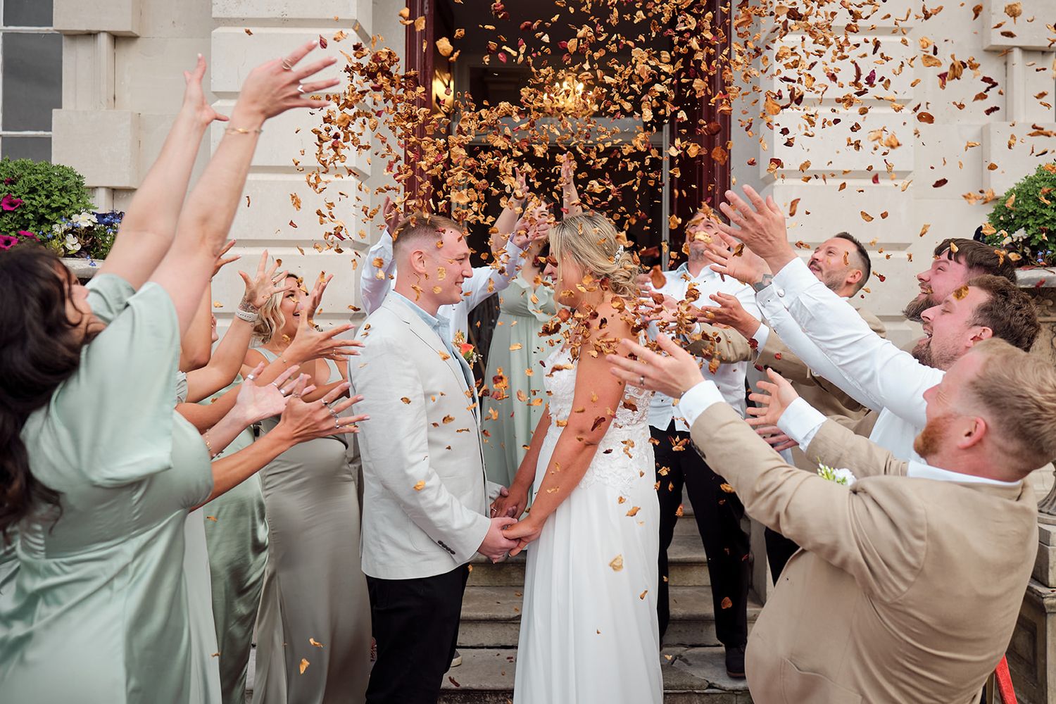 Wedding party throws autumn leaves in celebration outside elegant building during joyful ceremony moment.