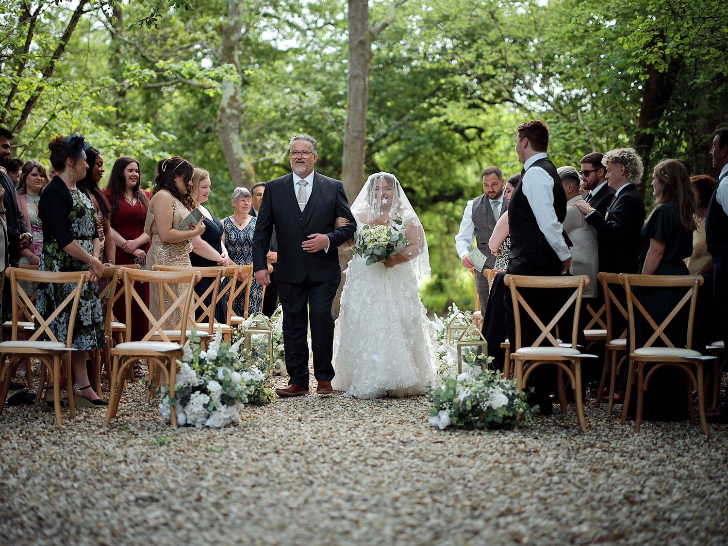 Wedding guests seated in rustic wooden chairs watch a romantic outdoor ceremony under a canopy of green trees at Rudge Farm Cottages.