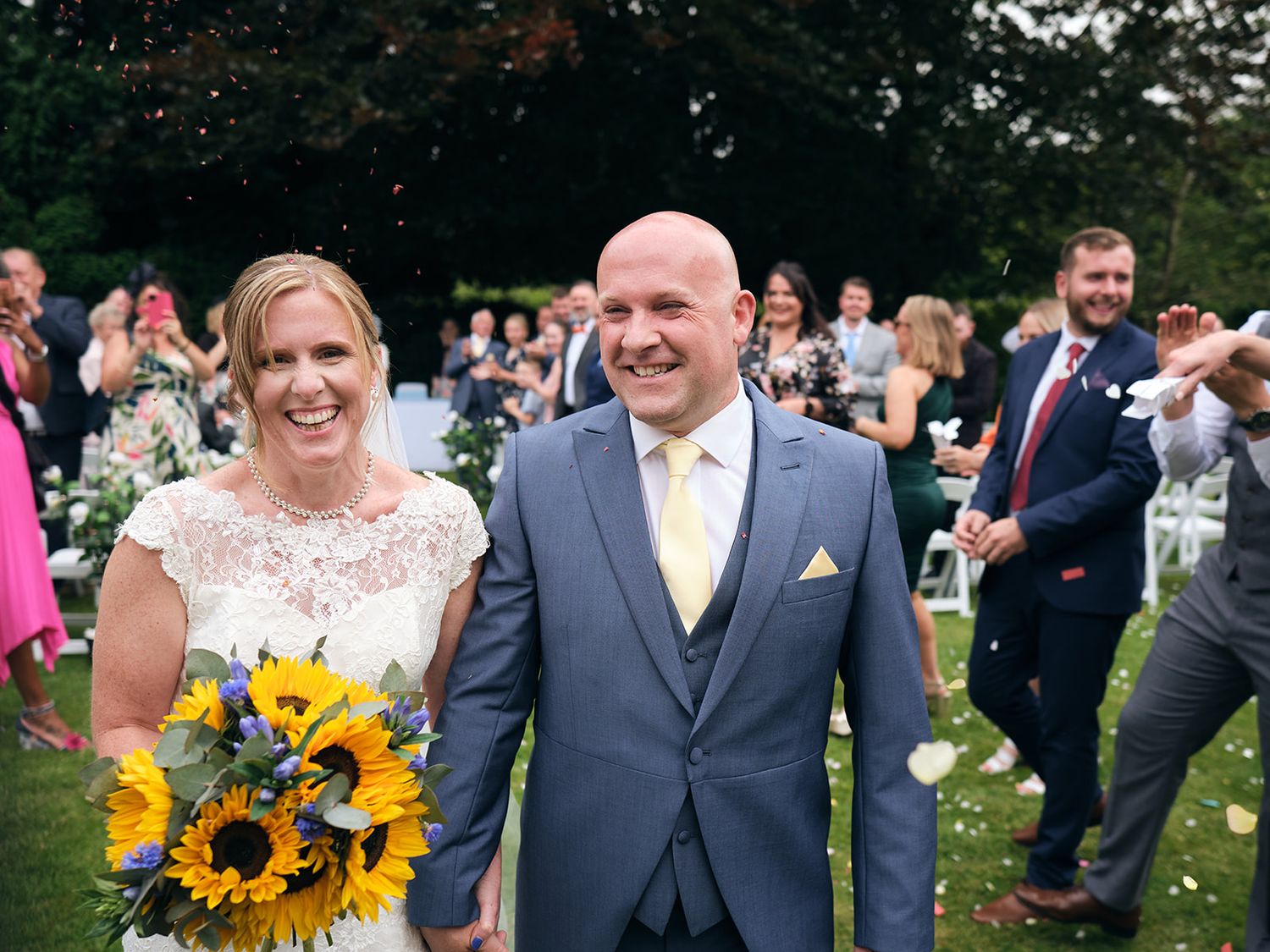 Wedding couple shares a joyful moment outdoors with guests as bride holds vibrant sunflower bouquet.