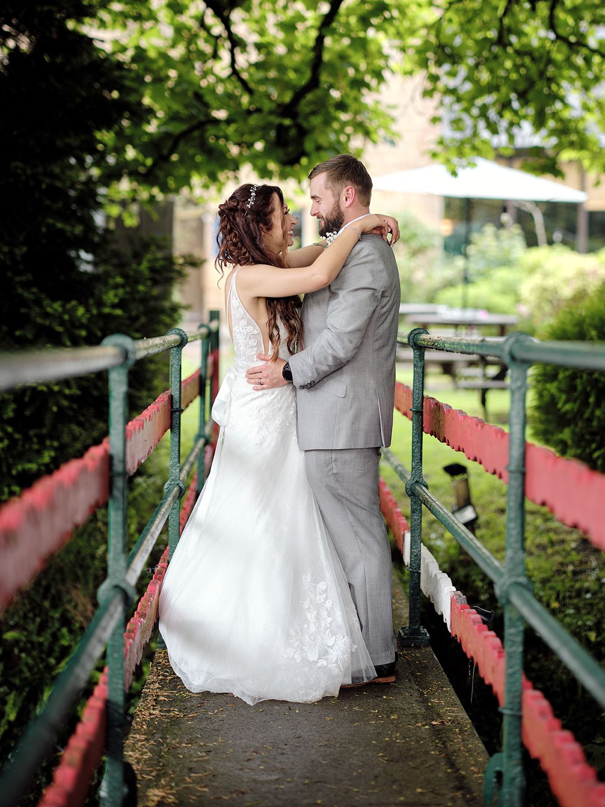A romantic wedding couple embraces on a red and green footbridge surrounded by lush summer foliage.