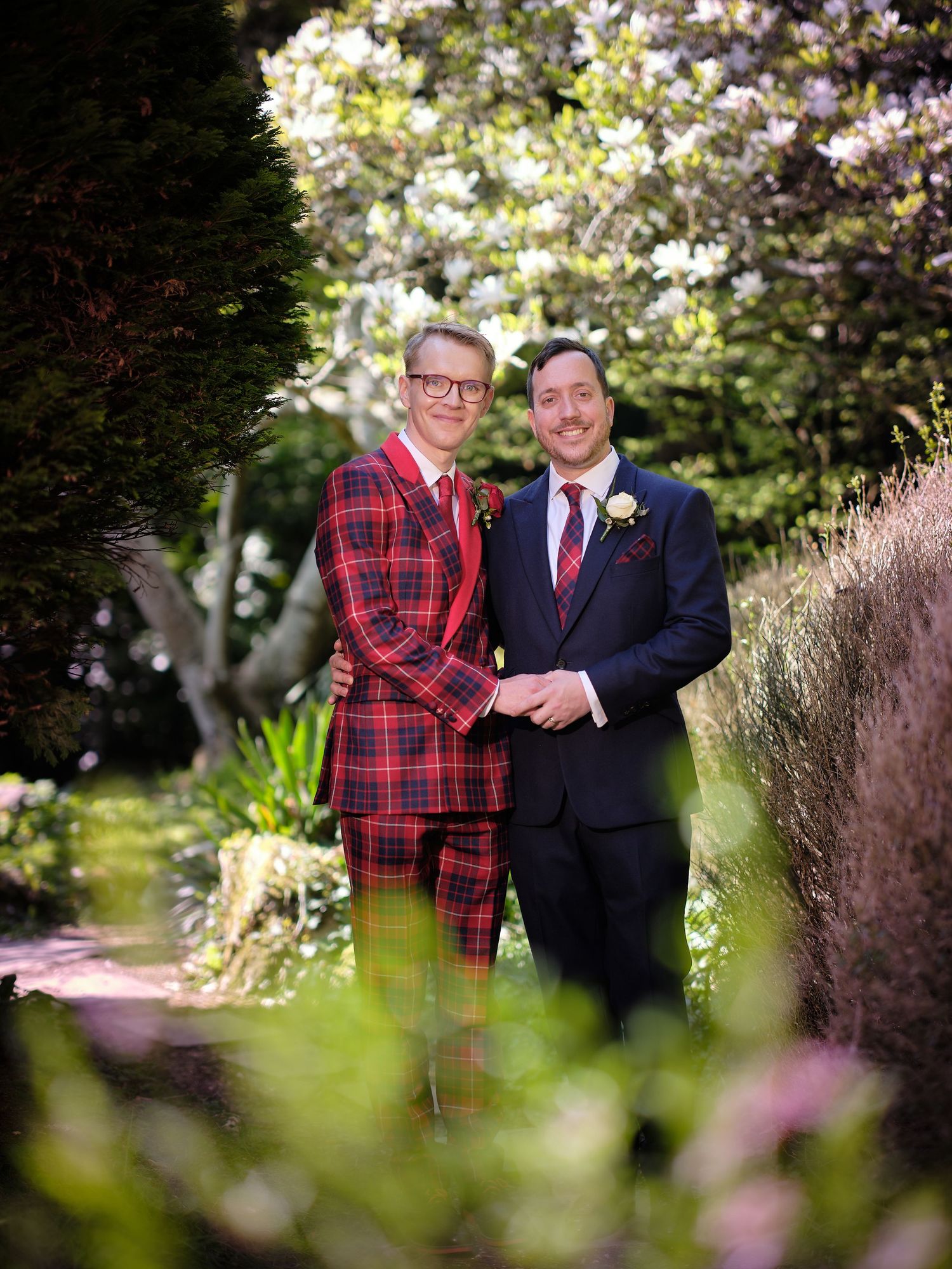 Couple in contrasting outfits - one in a bold red plaid suit, one in navy - pose in a blooming garden setting.