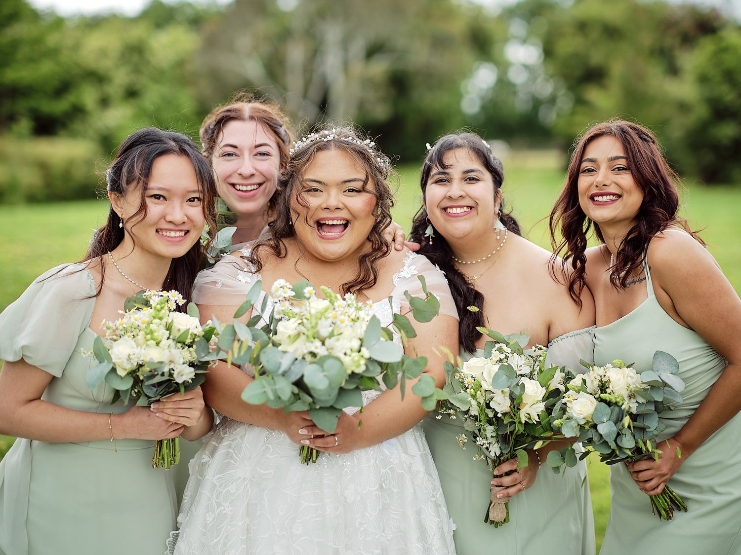 Bridesmaids in sage green dresses stand close together with white bouquets and share joyful laughter outdoors.