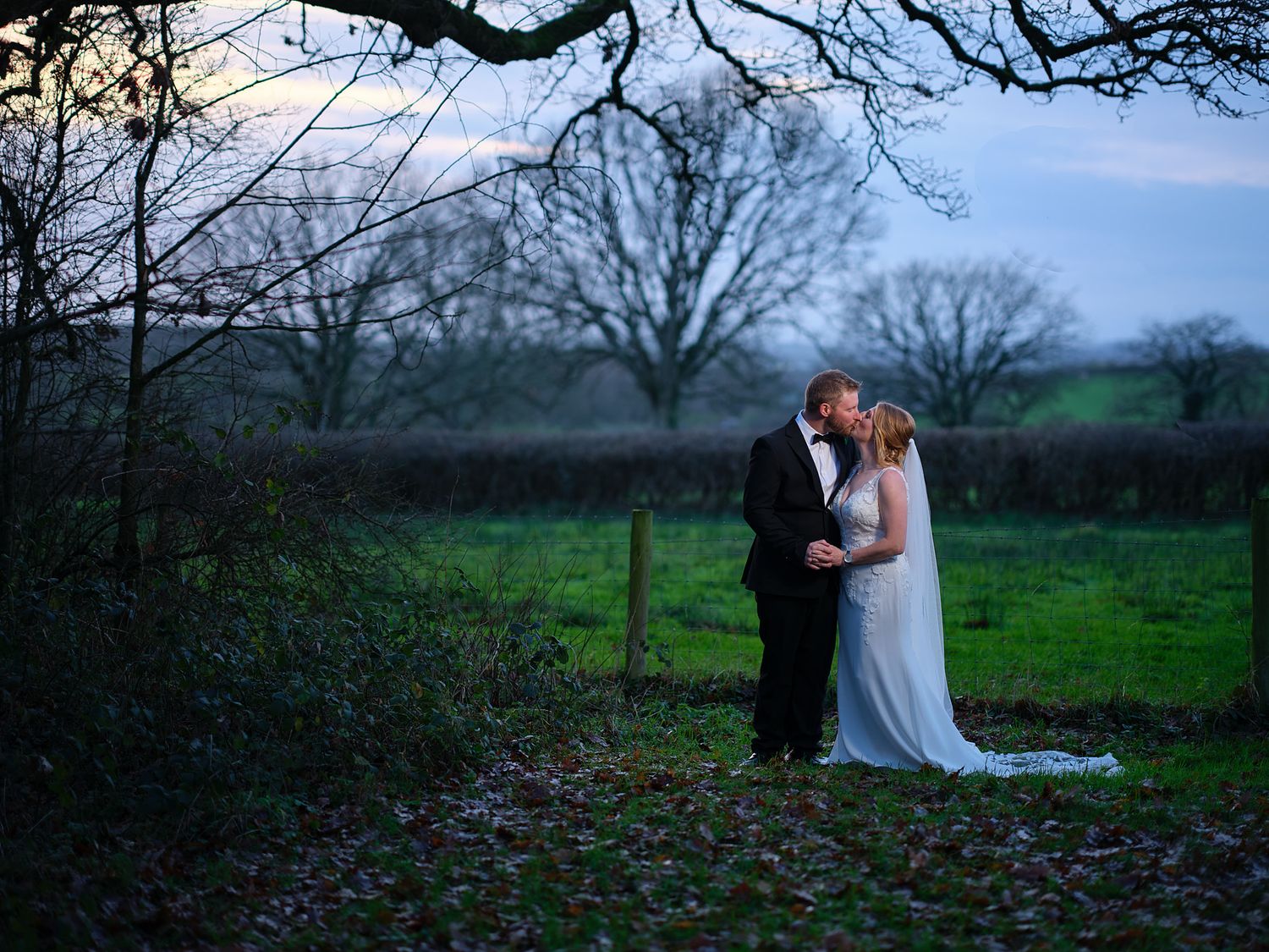 A couple shares a romantic moment at dusk in a misty countryside setting framed by bare winter trees and green fields.