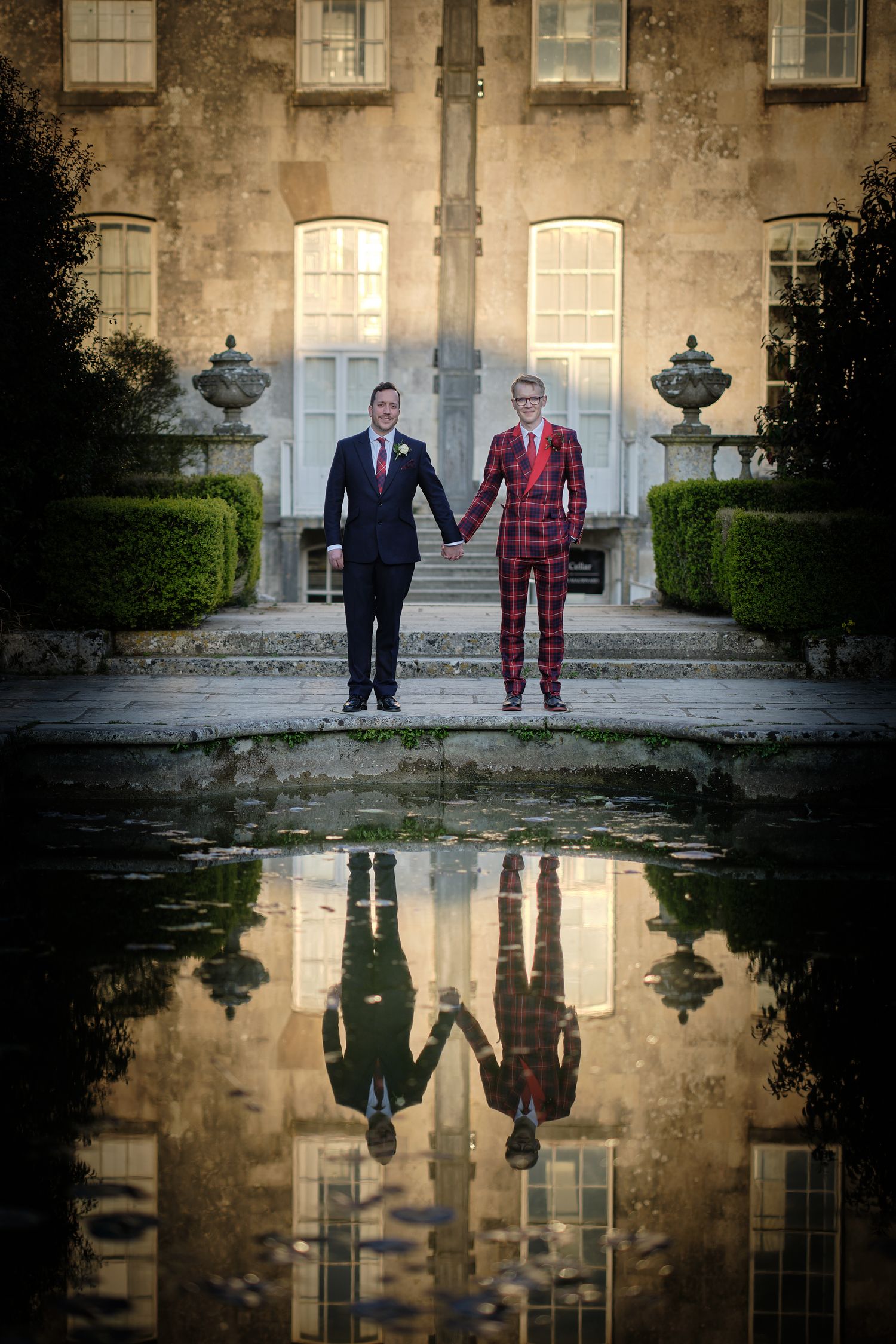 Groom and groom in formal near a reflecting pool outside Kingston Maurward with their reflection mirrored in the water.