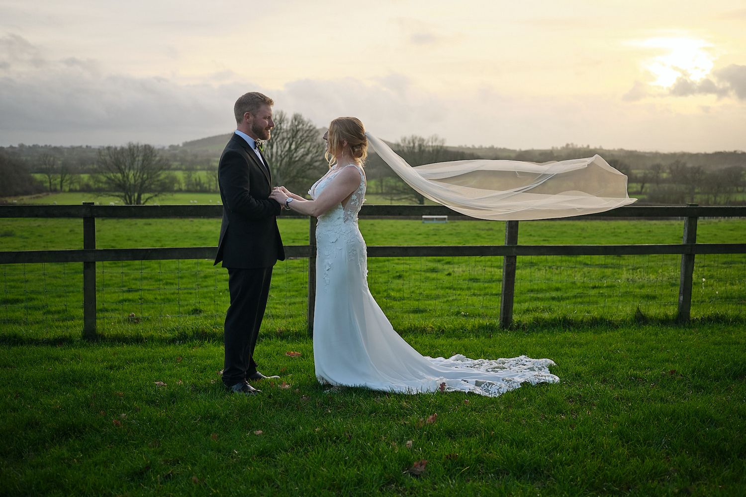 A couple in wedding attire stands in a green field with flowing veil at sunset creating a romantic silhouette.