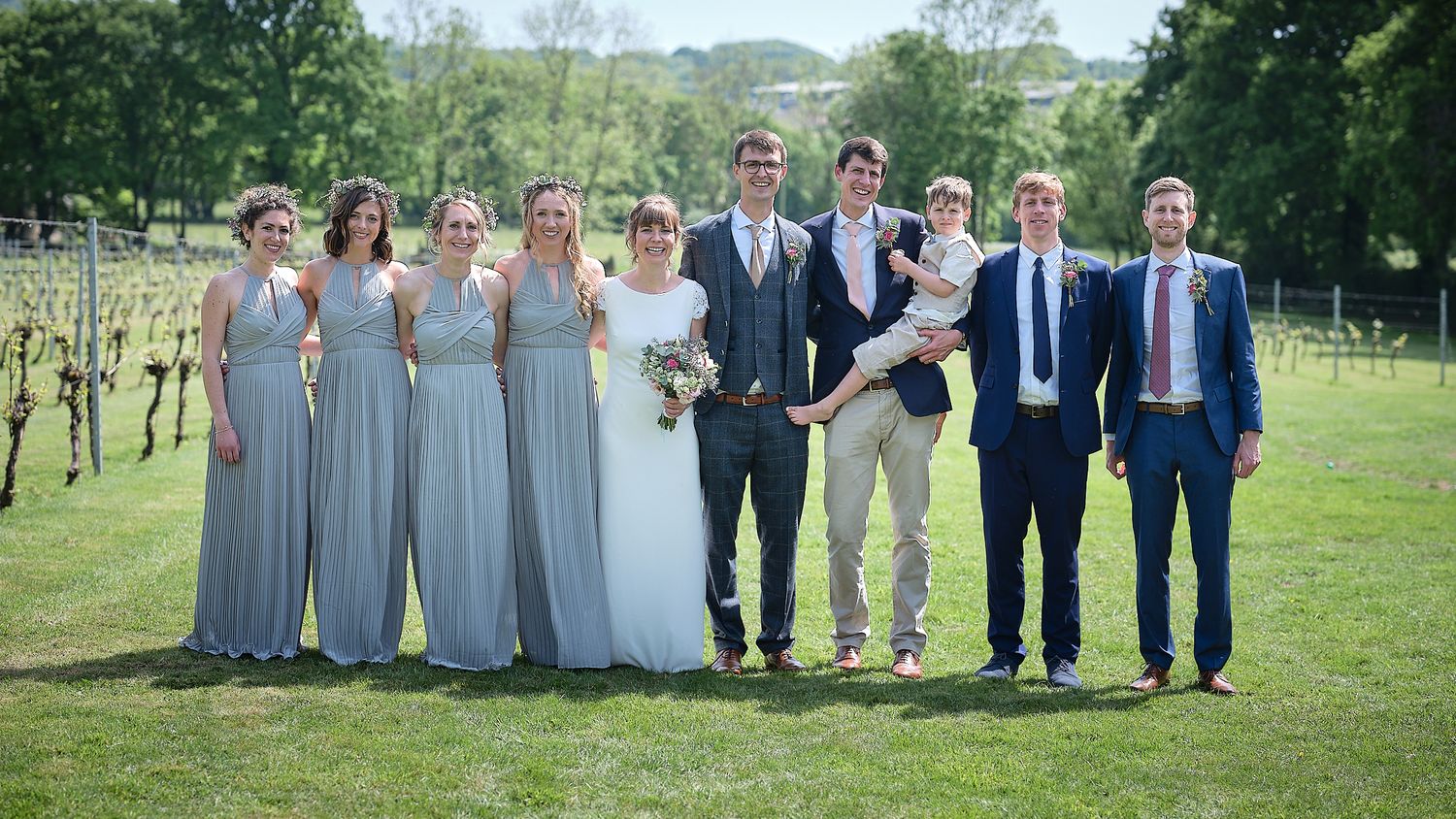 Wedding party stands in line on sunny day in front of vineyard wearing grey and blue formal attire.