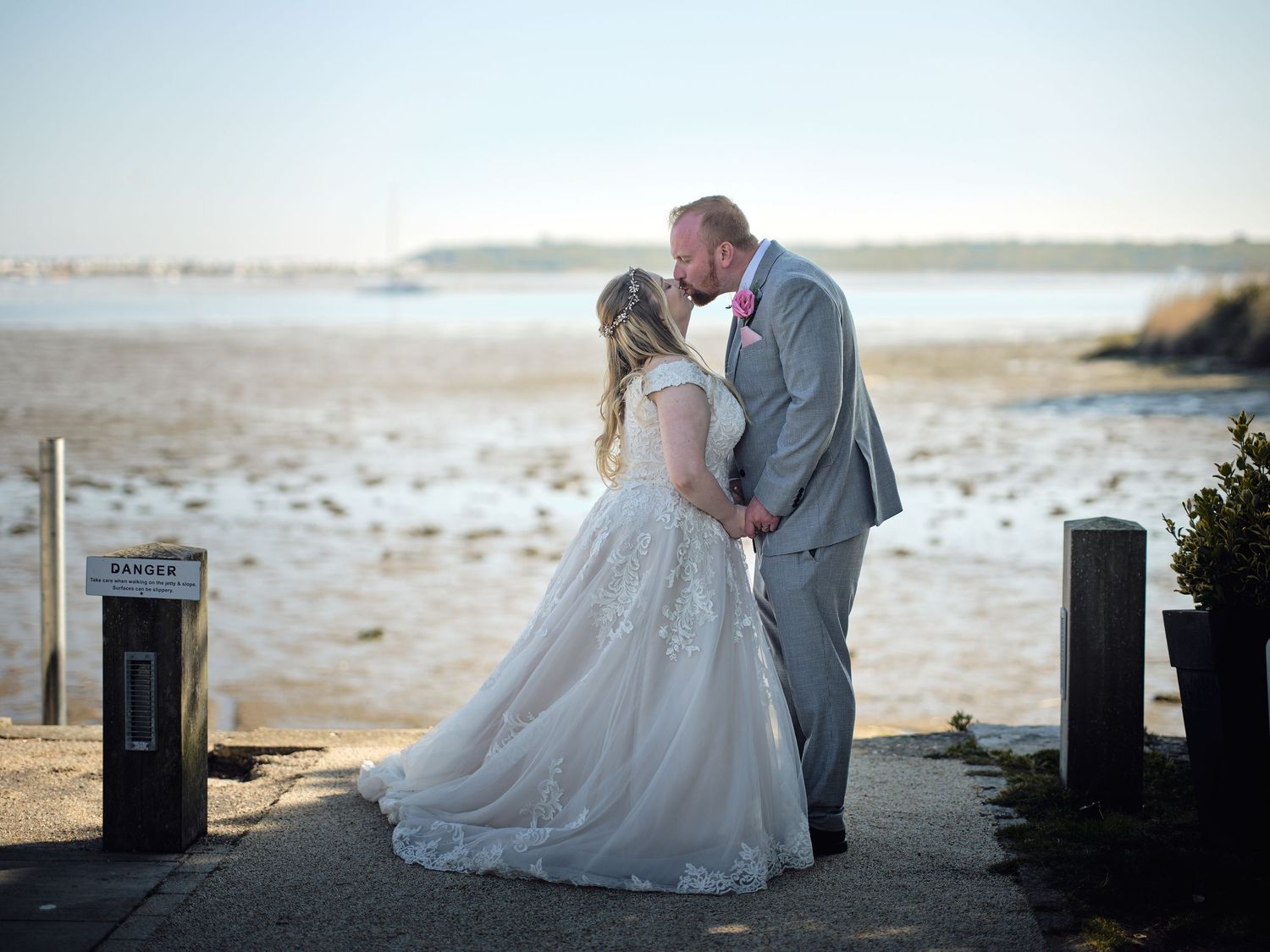 A couple shares a romantic kiss on a beach at sunset, with the bride wearing a flowing white lace gown and groom in gray suit.
