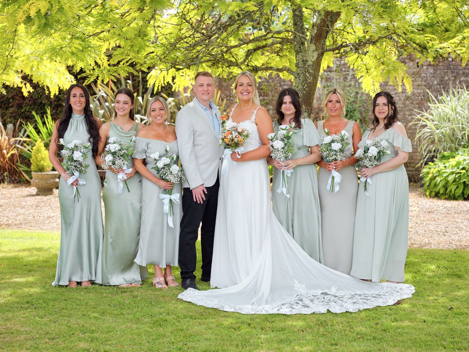 A wedding party outdoors in a lush garden with the wedding party wearing sage green bridesmaid dresses.