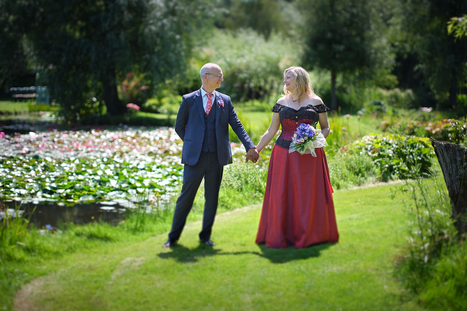 A couple stands together on a green lawn near a colorful flower garden on a sunny day at Bennetts Water Gardens.
