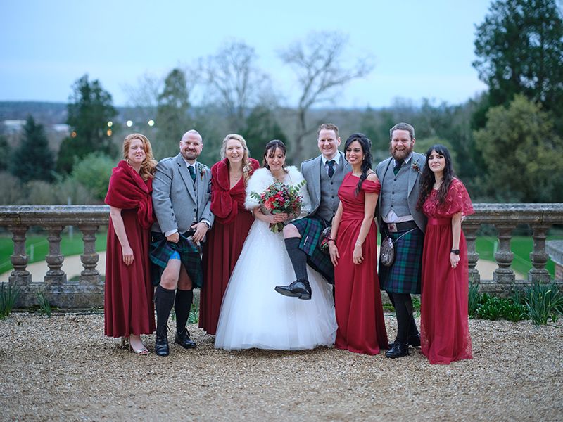 Wedding party in burgundy bridesmaid dresses poses together outdoors for formal photos in a garden setting.