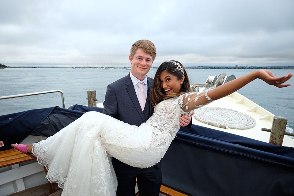 A newlywed couple poses together on a boat in Poole Bay with cloudy sky and waterfront in the background.