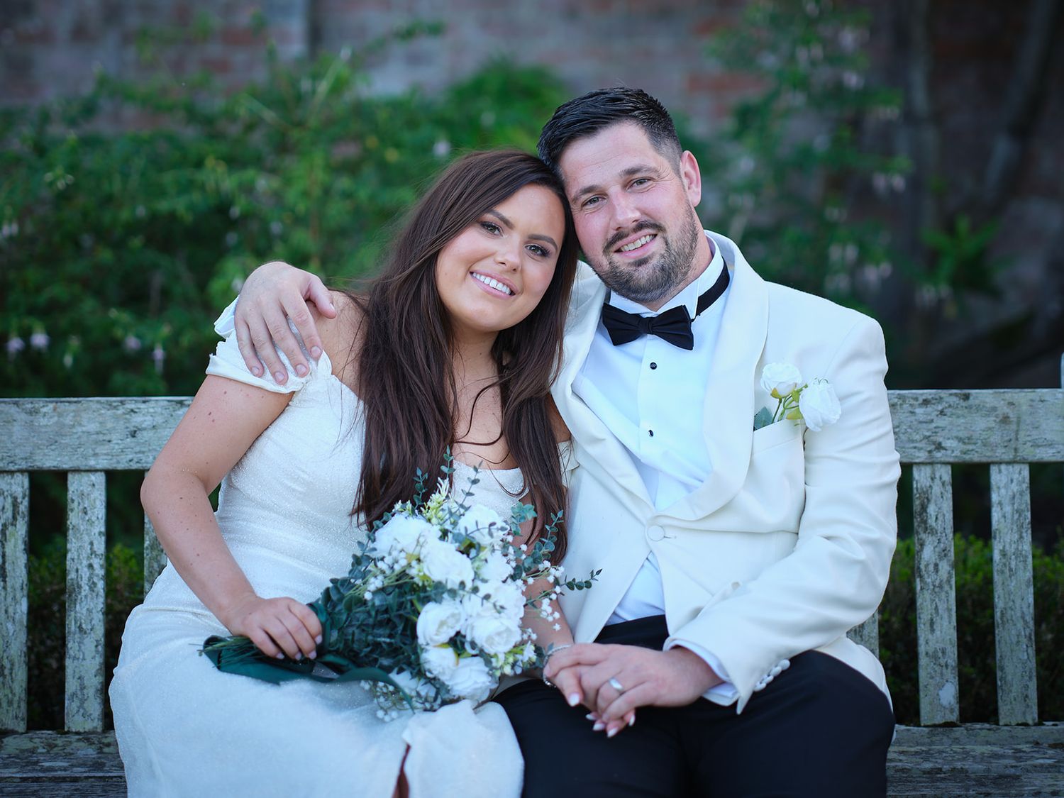 A bride and groom share romantic moments together on a wooden bench in an outdoor garden setting.
