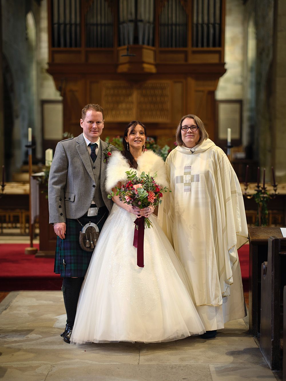 A wedding couple and clergy member walk through a traditional church interior with stone columns and wooden pews.