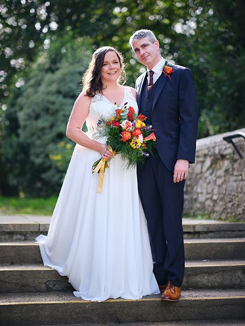 Wedding couple poses on stone steps outdoors with bright autumn-colored bouquet in front of trees and bokeh lighting.
