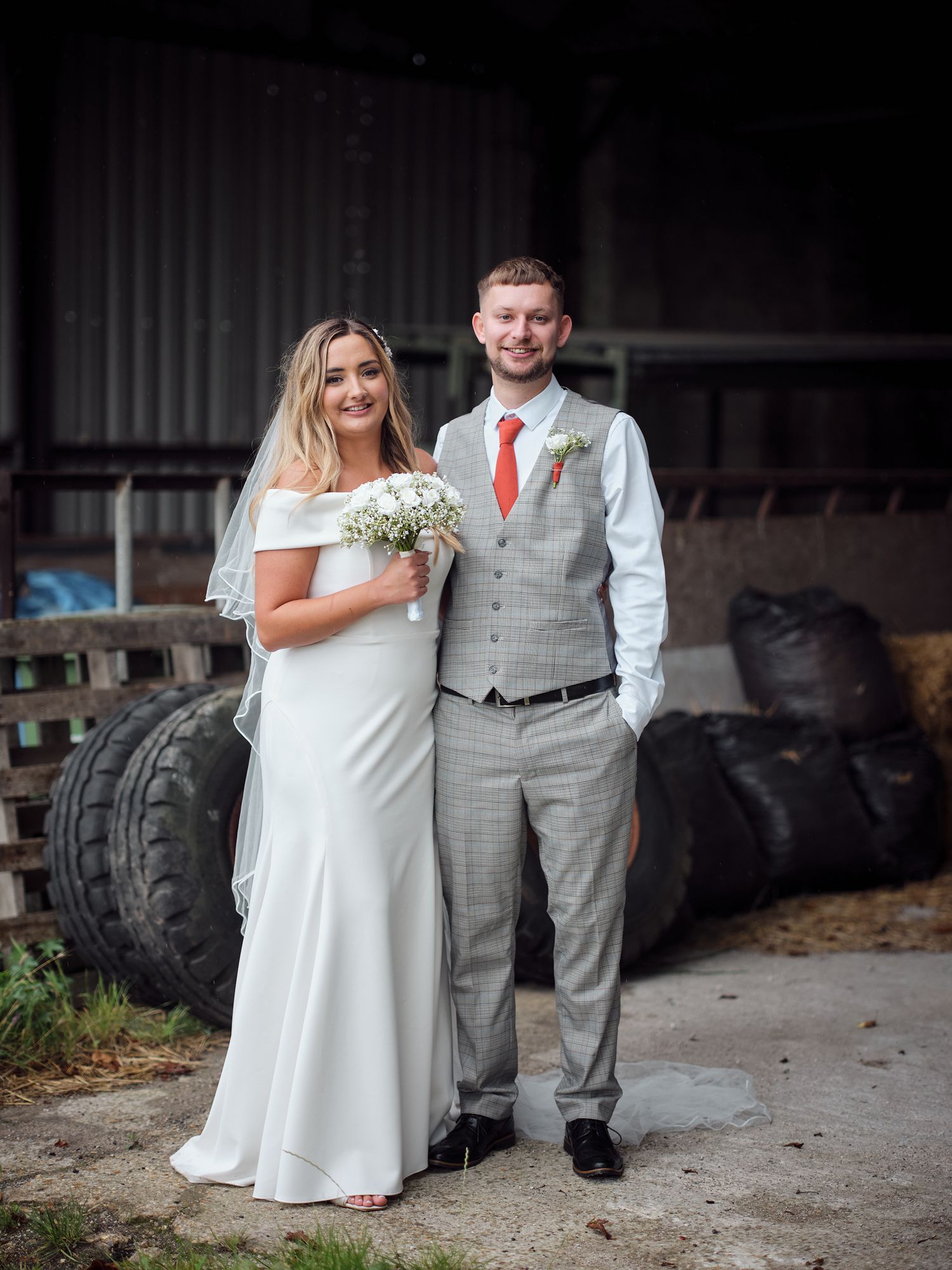 A couple poses in their wedding attire in front of a rustic barn setting with farm equipment and tires in the background.
