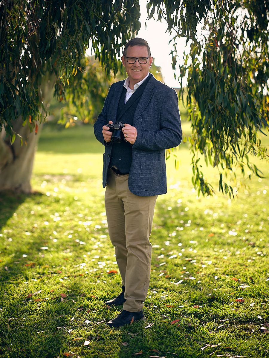Wedding photographer Ed Coleman in a navy blazer and khakis stands under a willow tree on a sunny autumn day.