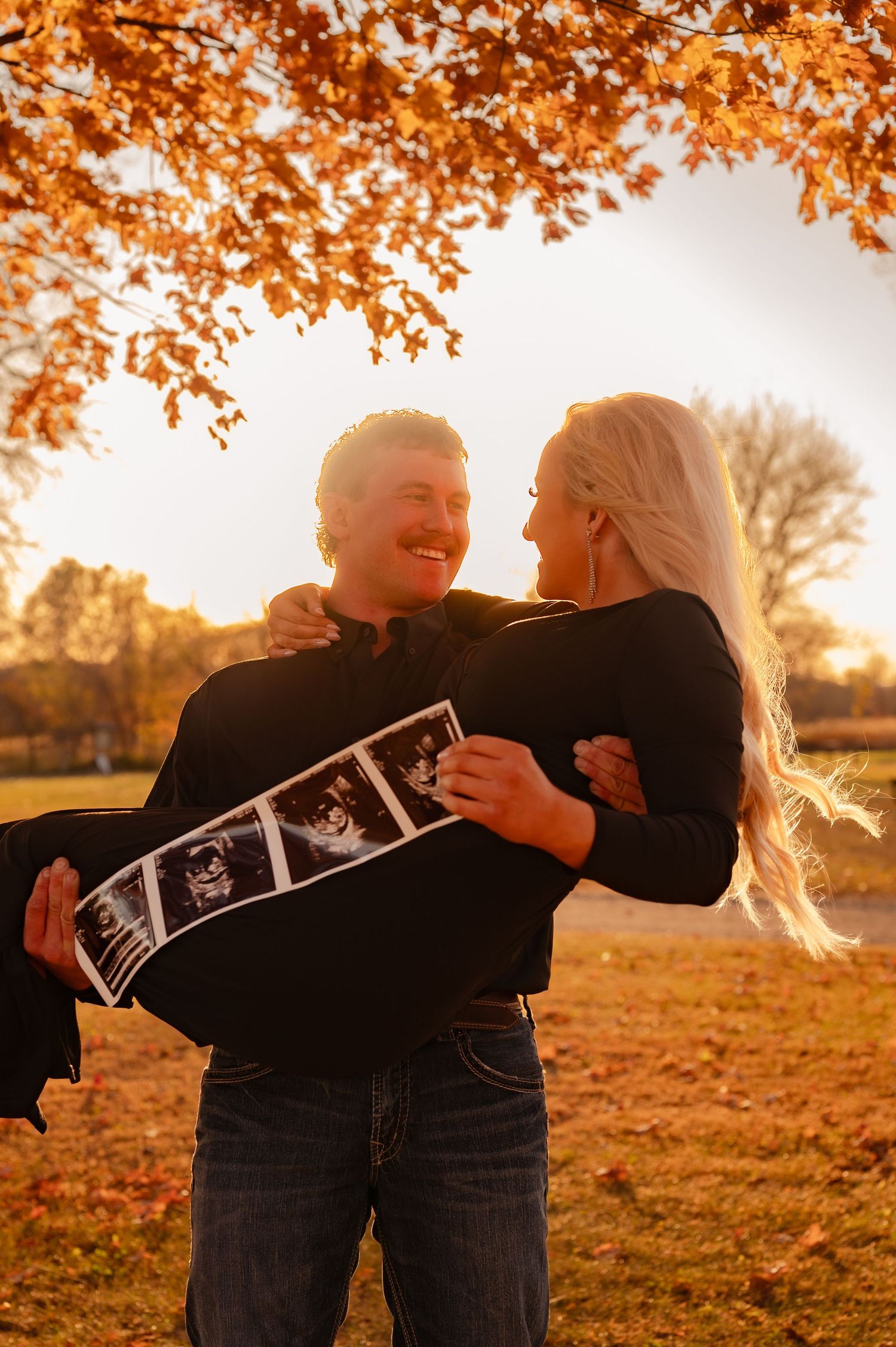 A couple shares a joyful moment under autumn trees while holding ultrasound photos during a pregnancy announcement photoshoot.