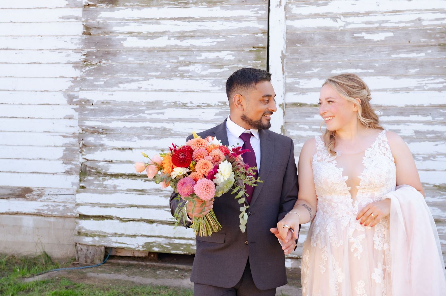 A couple shares romantic moments in their wedding attire against a rustic white wooden wall background.