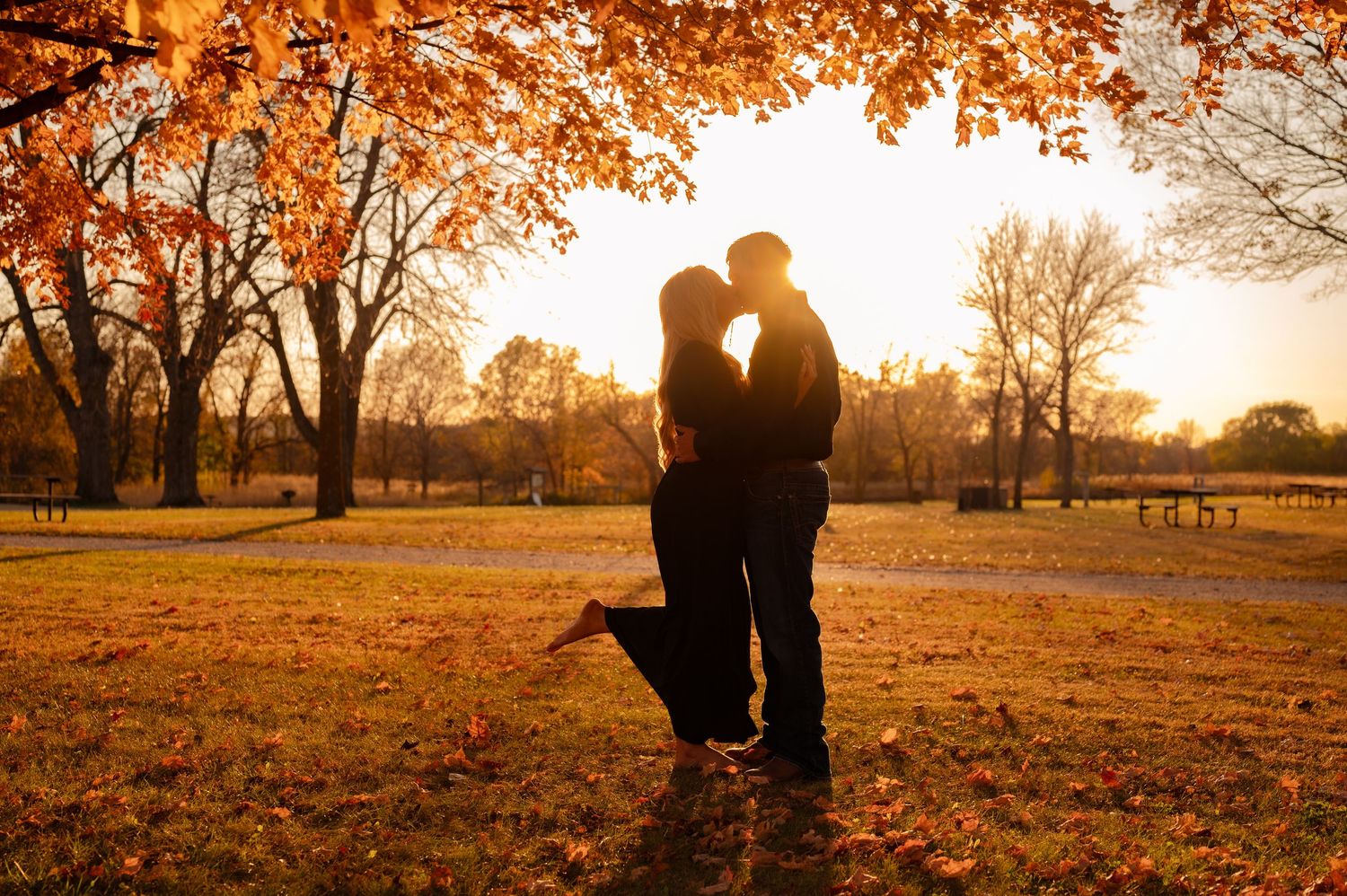 Silhouettes embrace during golden sunset in autumn park with orange foliage.