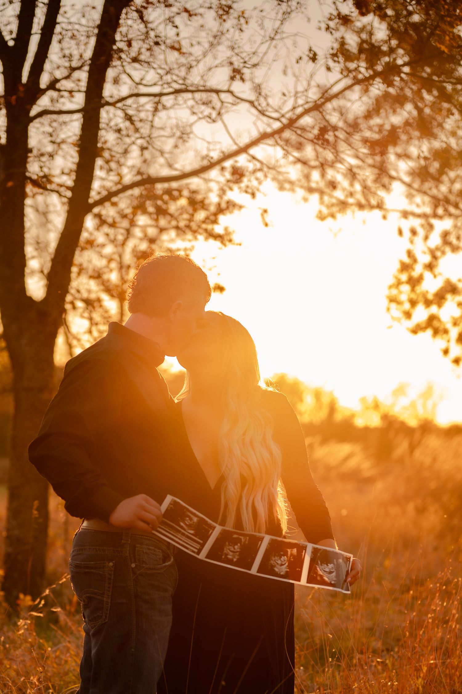 Silhouetted couple embracing in a field during sunset with trees creating a dreamy autumn backdrop.