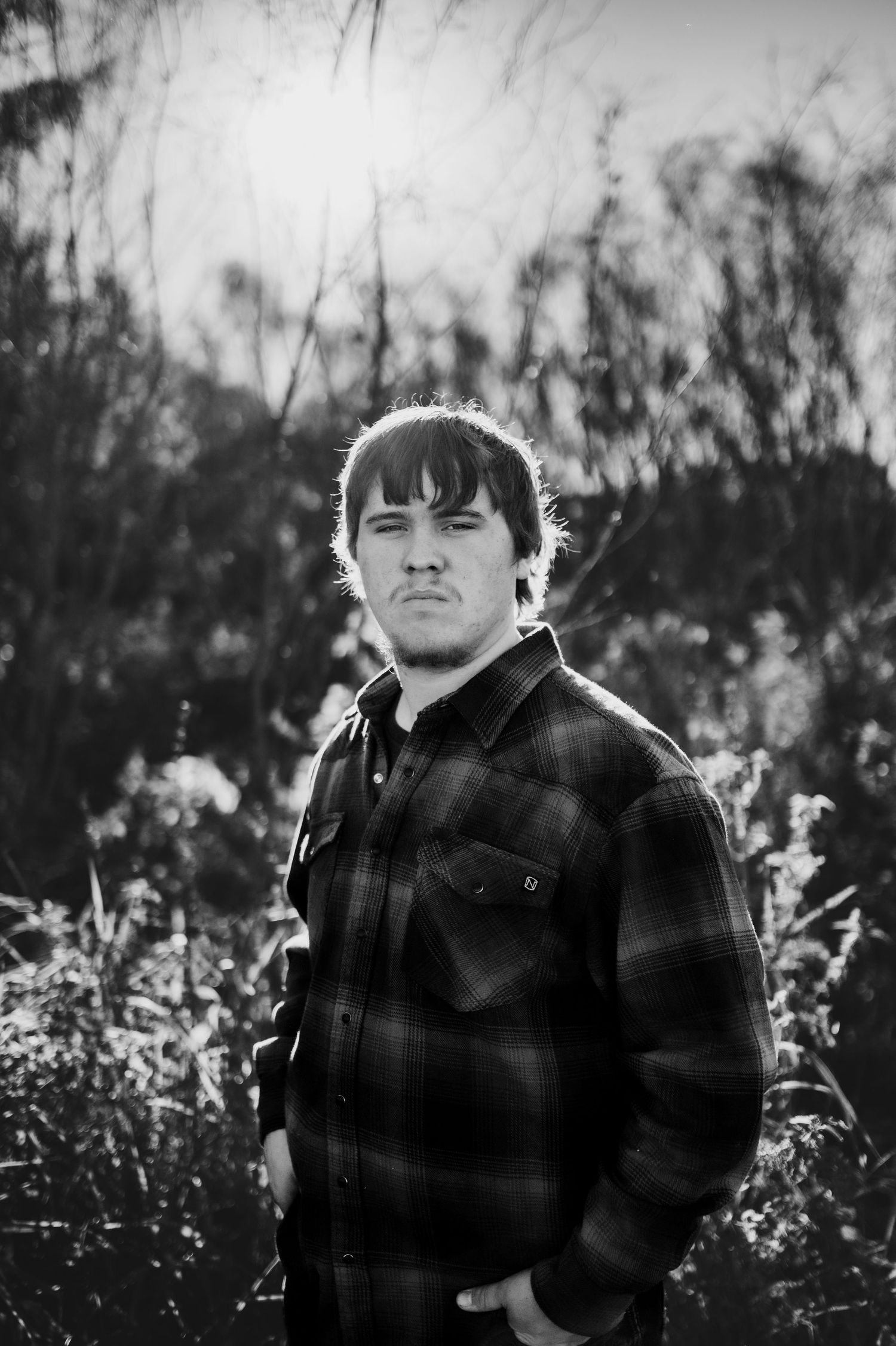 Black and white portrait series showing person in plaid jacket outdoors with dramatic backlit glow through trees.