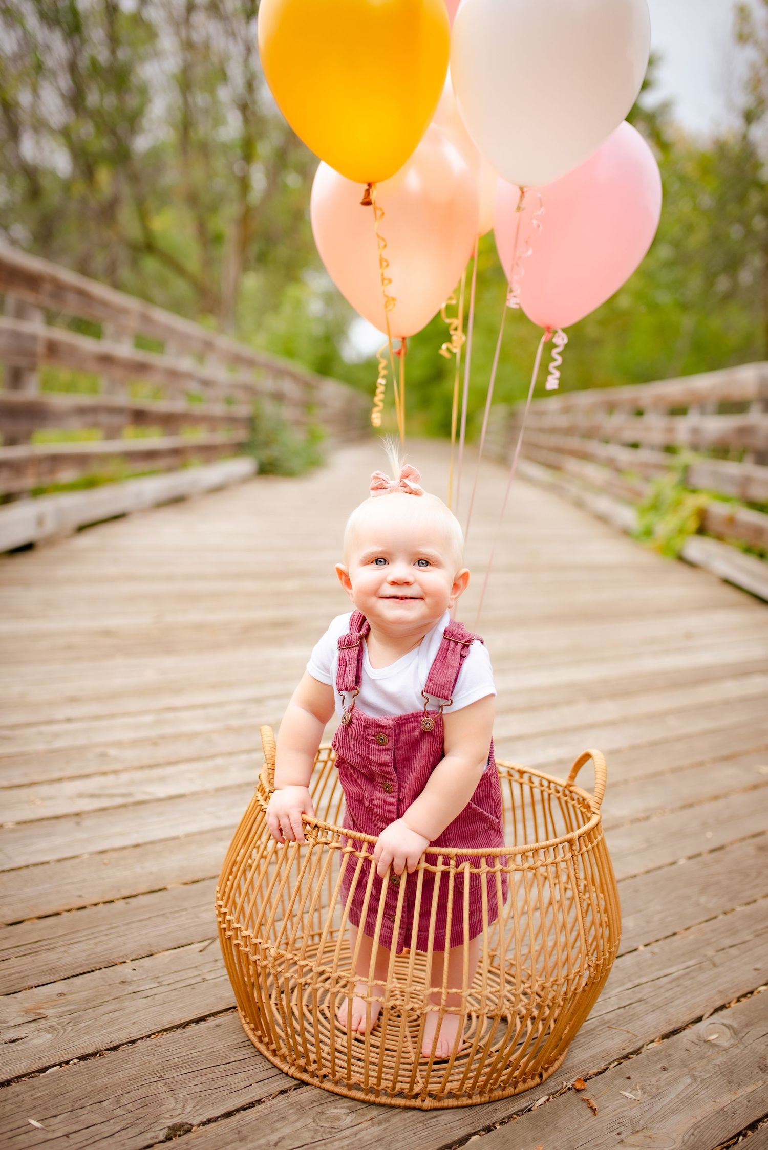 Baby in red overalls sits in wicker basket with colorful balloons on wooden bridge.