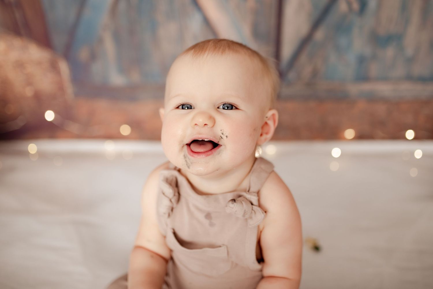 Smiling baby in beige outfit against rustic backdrop with twinkling lights.