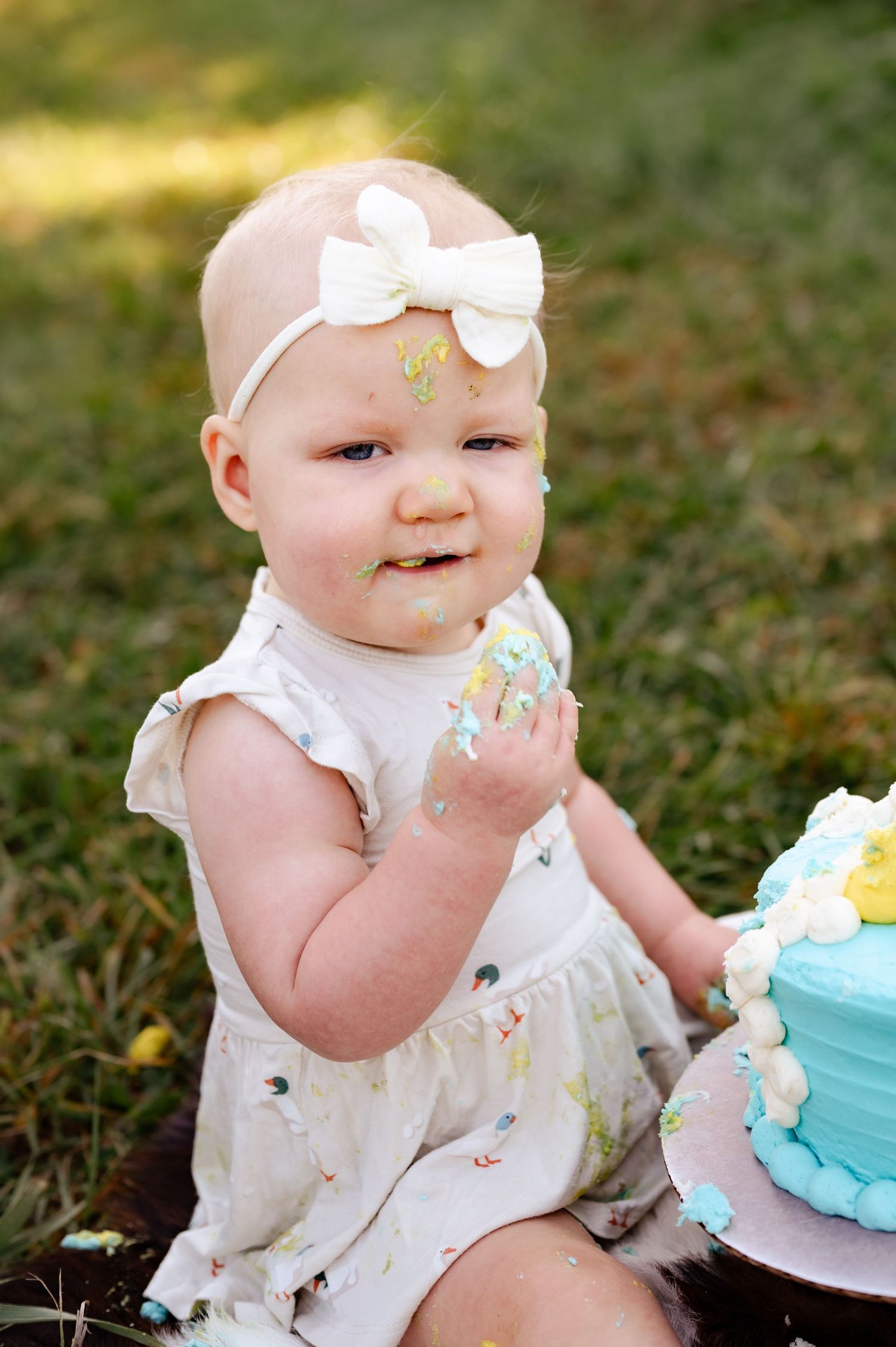 Baby in white dress enjoys first birthday cake smash photoshoot outdoors.