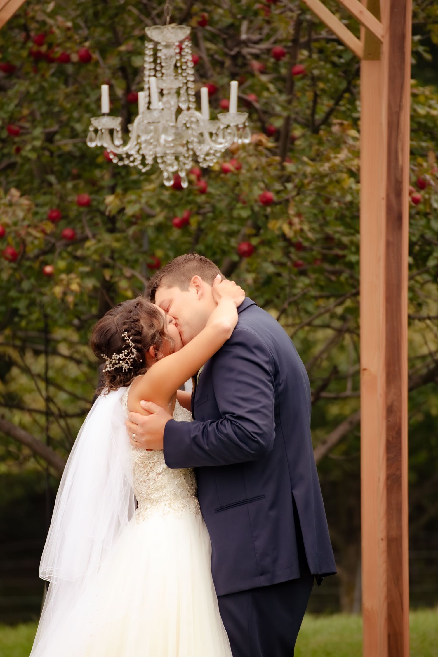 Romantic wedding kiss under an elegant crystal chandelier hanging from a wooden ceremony arch.