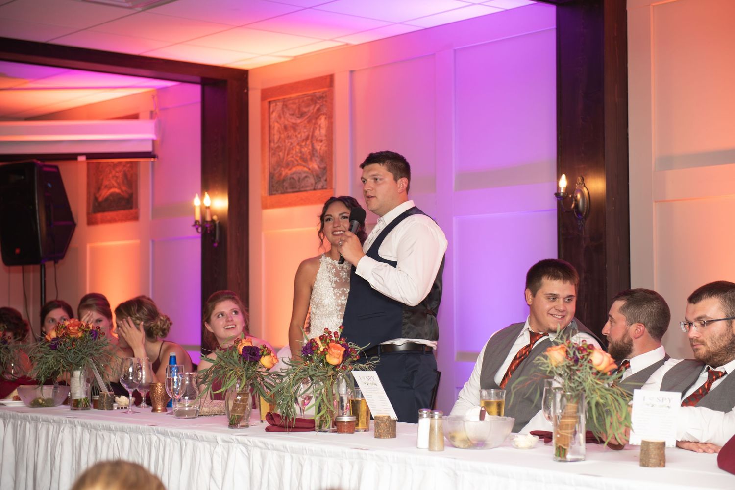 Wedding party seated at decorated head table during reception toasts and speeches.
