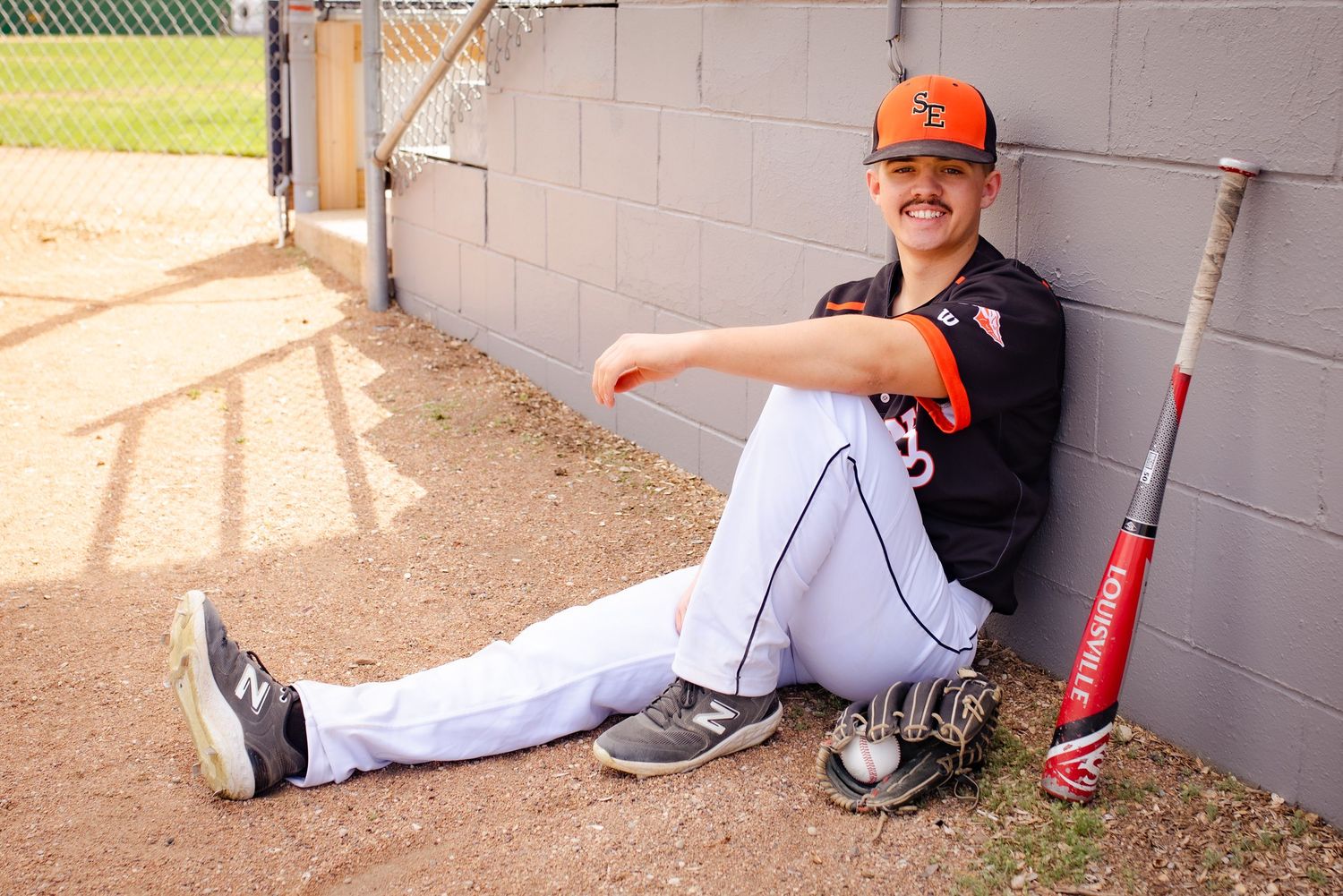 Baseball player in uniform sits against wall in dugout with bat beside him.