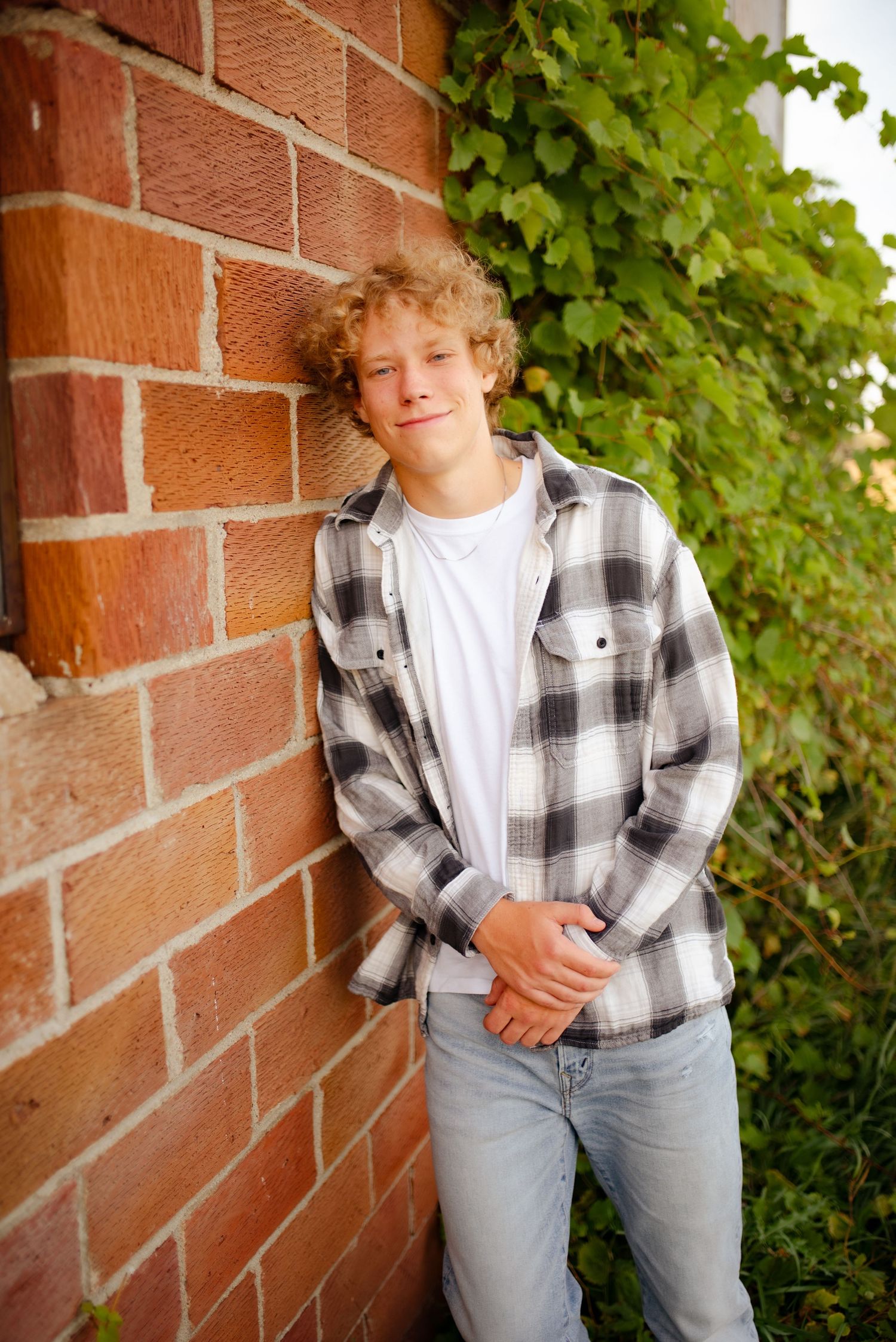 Portrait in front of brick wall with ivy, wearing plaid shirt and jeans in warm lighting.