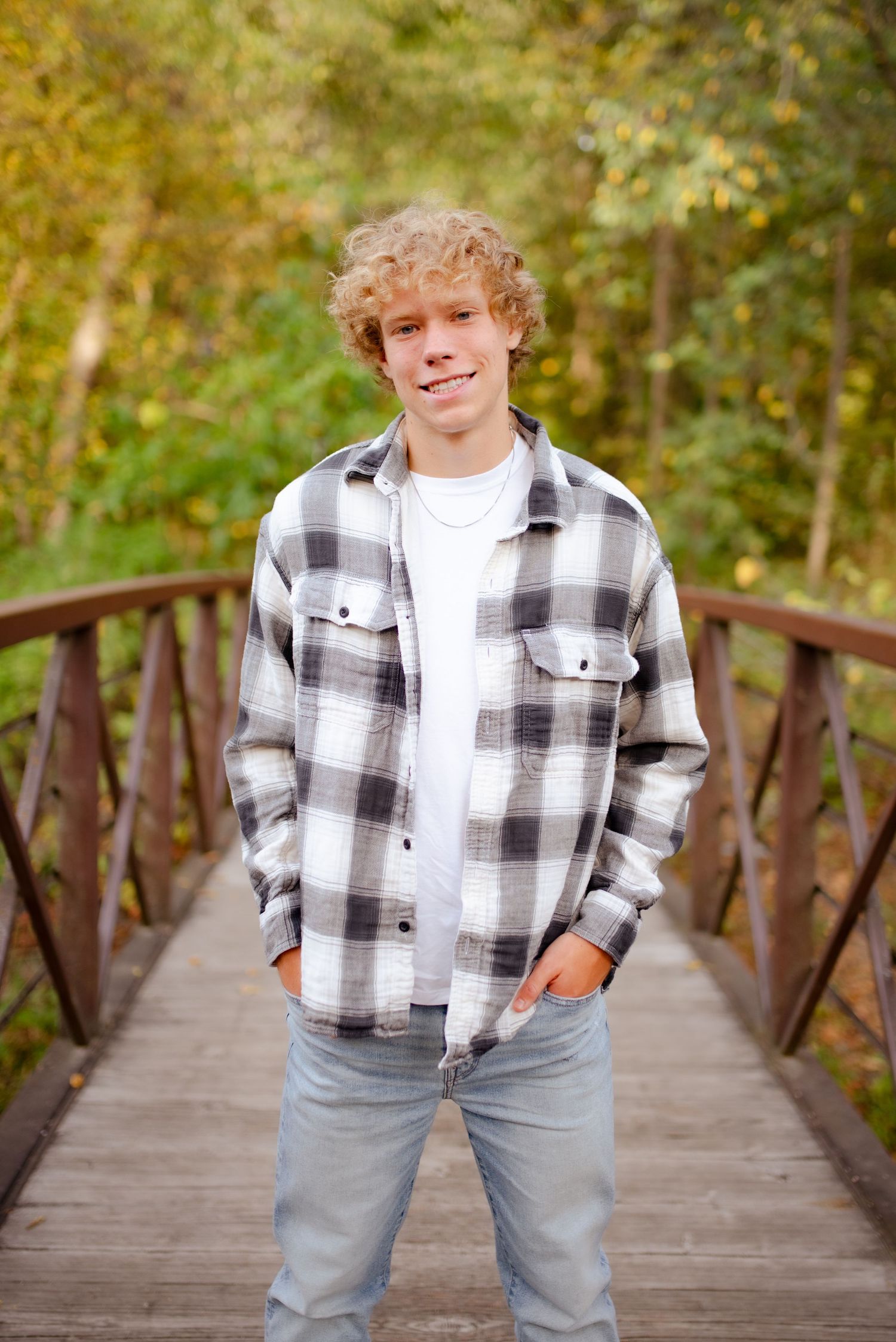A portrait of someone wearing a plaid flannel shirt on a wooden bridge with autumn foliage.