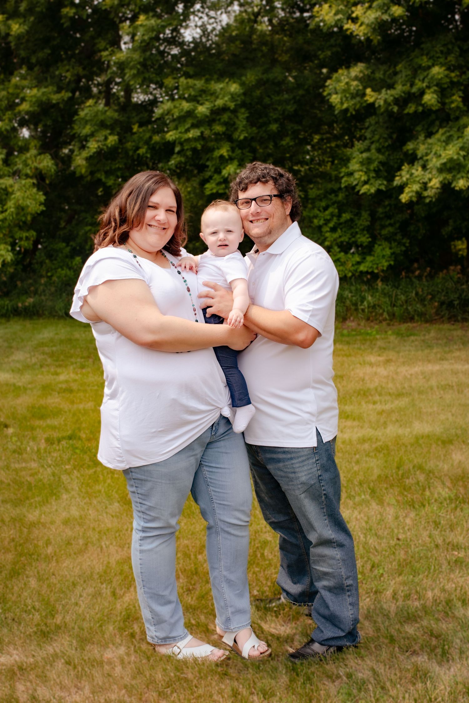 Family of three poses together in casual white outfits against green summer foliage.