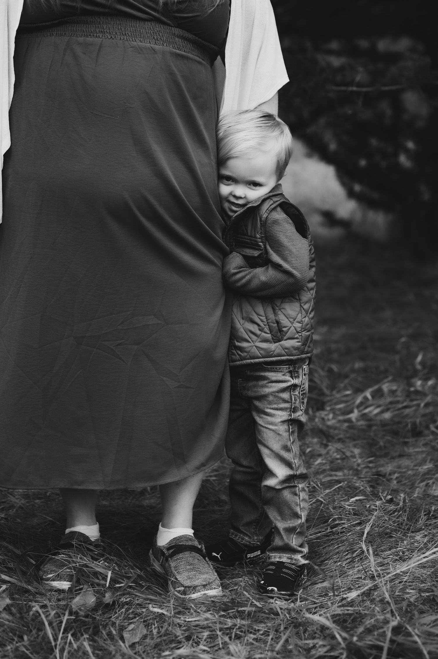 Black and white photograph showing small child's legs peeking out from behind an adult wearing long dark clothing.
